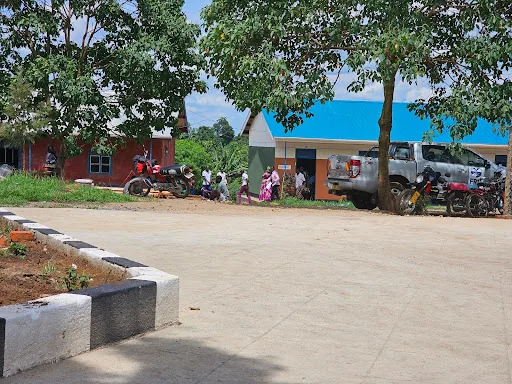 The school entrance yard with a paved area, trees, motorcycles and a pickup truck in front of the buildings.