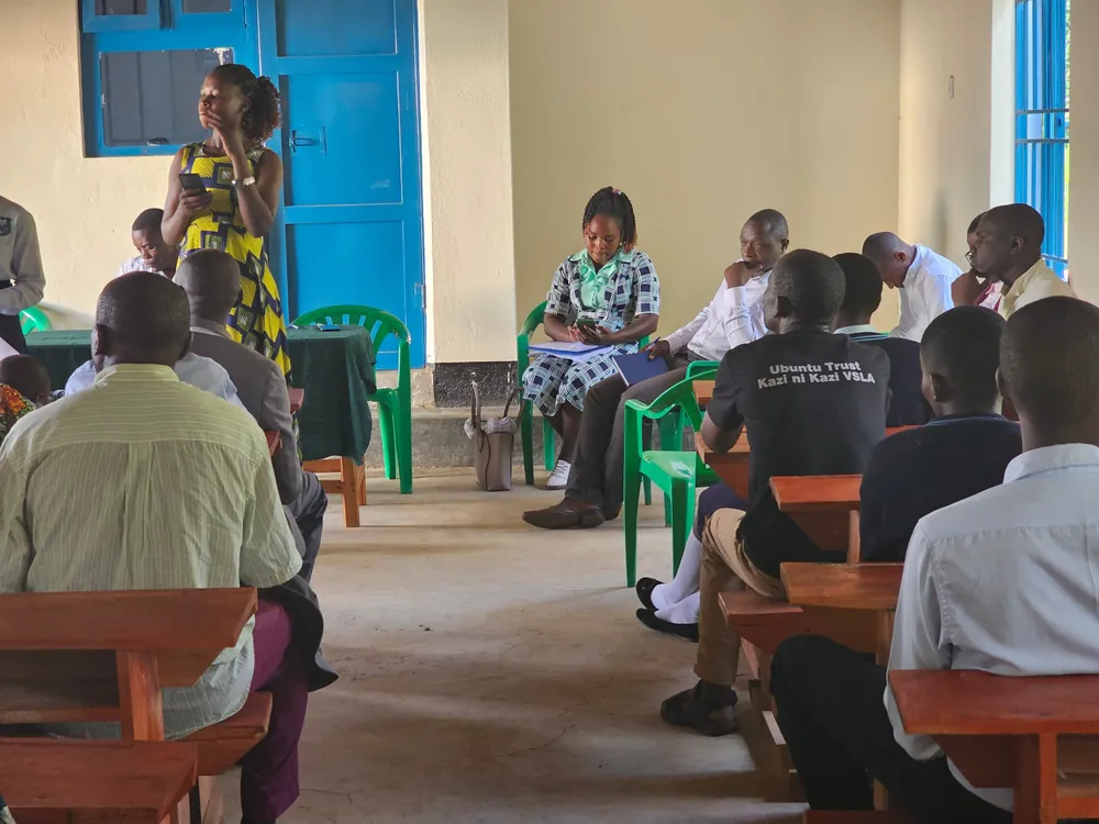 A facilitator stands and speaks to parents, staff and learners seated on benches and chairs in a school hall.