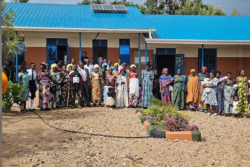 A large group of adults and some learners stands in front of a classroom block with a blue roof and solar panels, behind a small flower bed.