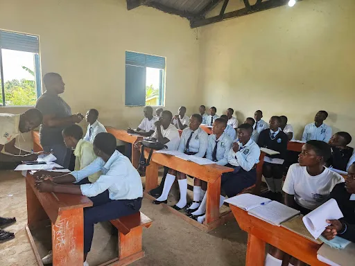 Learners in school uniform sit at wooden desks during a lesson, some reading, some listening.