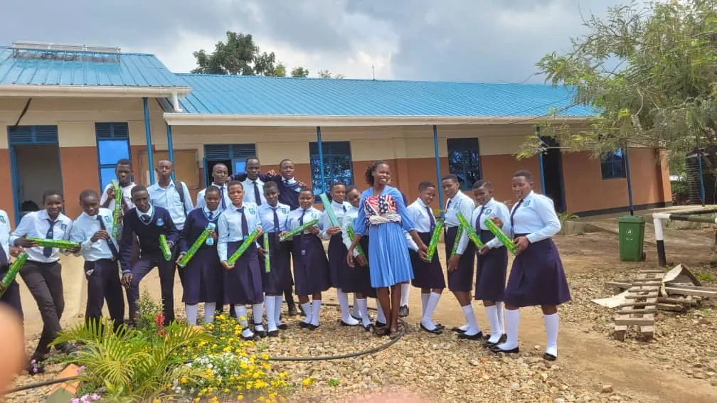 A group of learners in school uniform, each holding a green package, stands with a visitor in front of a classroom block.