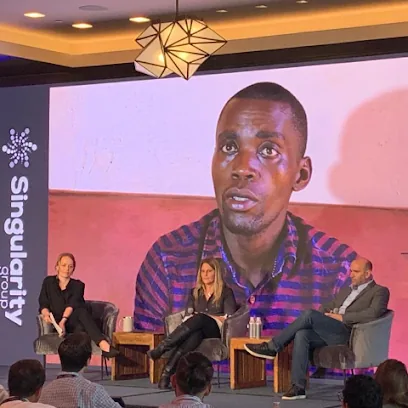 Brainy Swaibu shown on a large screen as he speaks on stage at a Singularity Group event, with three other speakers seated below.