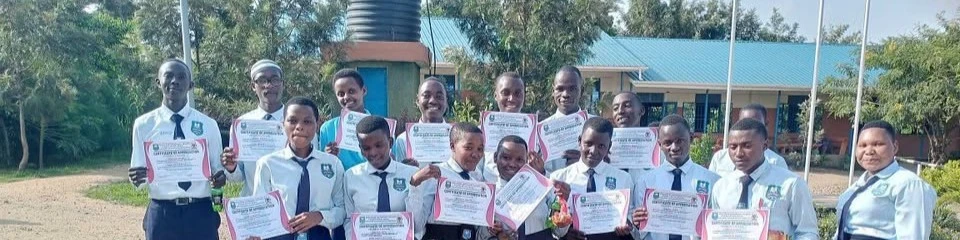 Learners in school uniform hold up certificates in front of a classroom block and water tower.
