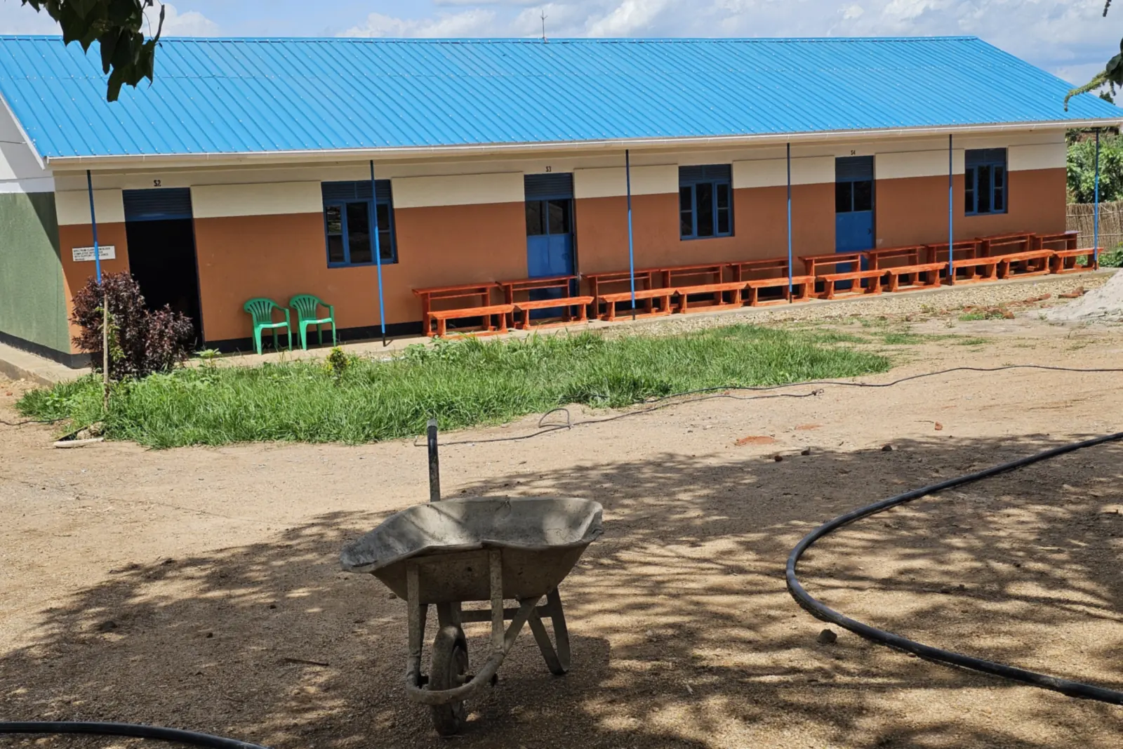 A long classroom block with a blue roof and blue doors, with a wheelbarrow in the shade in front of it.