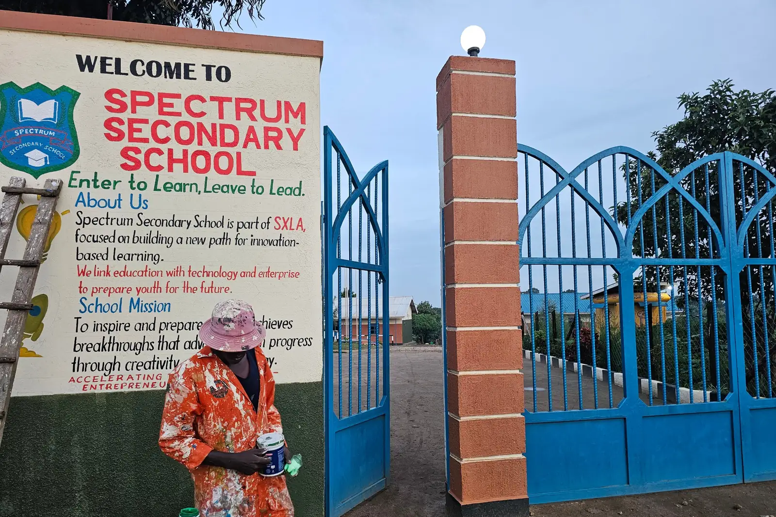 The new blue school gate beside a welcome mural that reads Enter to Learn, Leave to Lead, with a painter at work in front of the mural.
