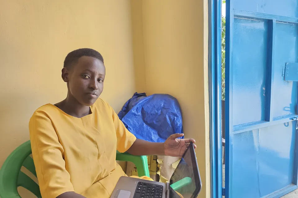 A secondary school learner in a yellow dress sits by a classroom door with a laptop open on her lap.
