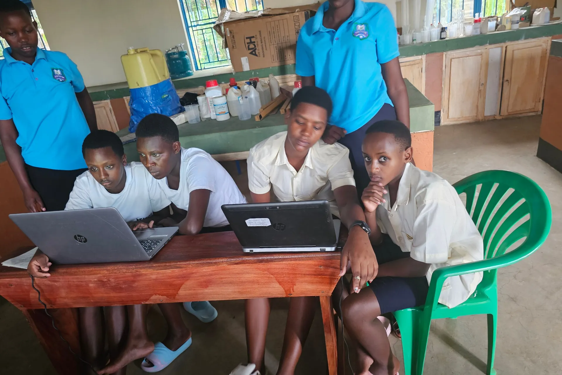 Girls in school uniform work together on two laptops at a table in the school laboratory, while two others look on.