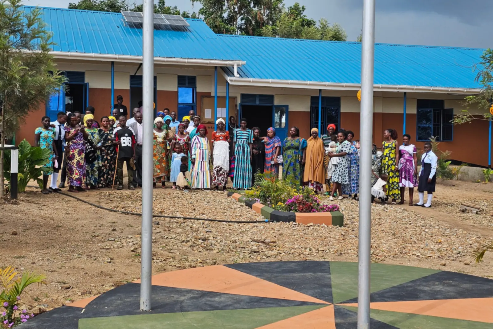 Parents and community members stand together in front of a blue-roofed classroom block, beside the school's flagpoles and painted flag stand.