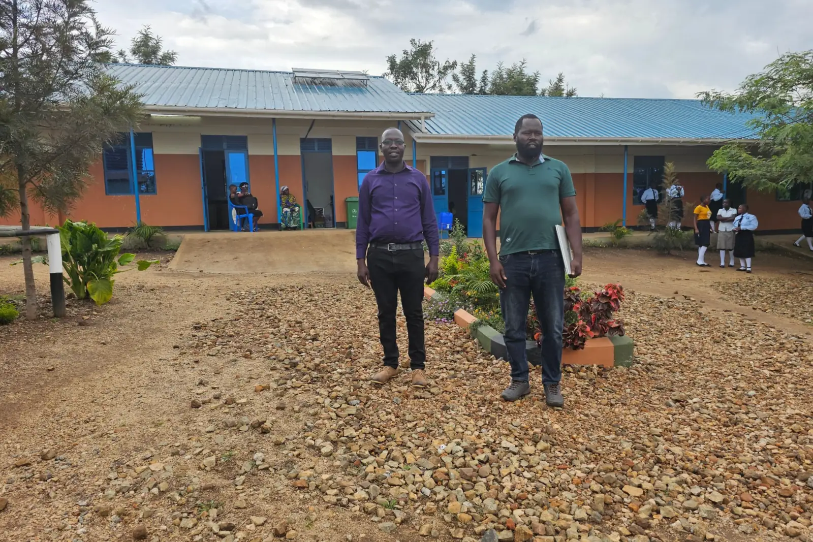 Two members of staff stand on the gravel in front of a classroom block, with learners in uniform in the background.